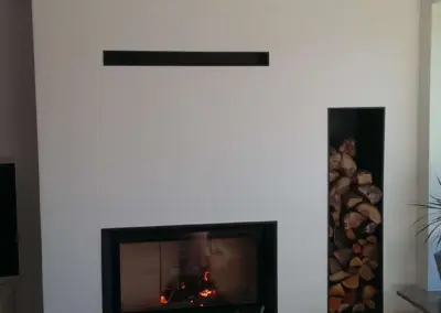 Moderner Heizkamin mit verputztem Aufbau, breiter Sichtscheibe und seitlich integriertem Holzfach im Wohnraum.