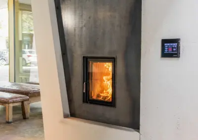 BRUNNER Kamin mit dunkler Verkleidung, hoher Sichtscheibe und integriertem Bedienelement an der Wand.