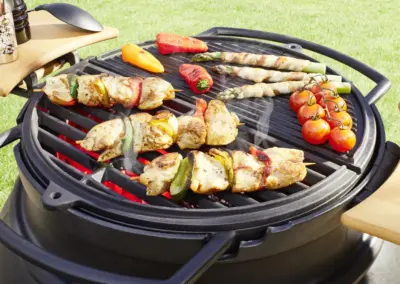 TROLL Gartenkamin mit Grillrost und Griddleplatte, belegt mit Fleischspießen, Spargel, Paprika und Tomaten.